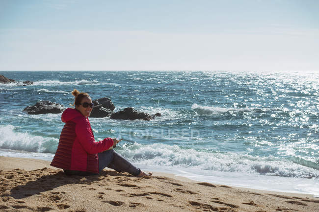 Зріла жінка сидить на березі моря в сонячний день і дивиться на камеру — стокове фото