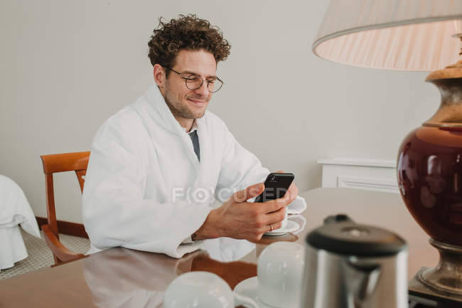 Mann im Bademantel surft mit Smartphone am Tisch — Stockfoto