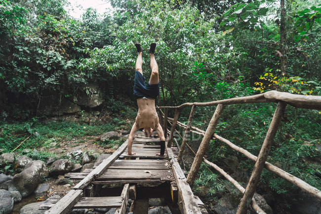 Hombre sin camisa de pie en las manos en el grungy puente de madera en el bosque verde
. - foto de stock