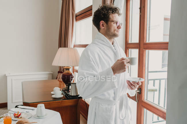 Человек в халате стоит с чашкой и смотрит в окно — стоковое фото