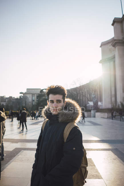 Joven pensativo de pie en la plaza de la ciudad bajo el sol y mirando hacia otro lado . - foto de stock