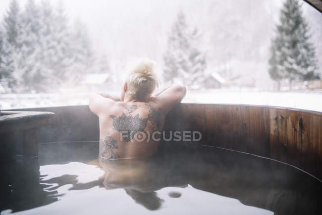 Back view of tattooed blonde woman swimming in plunge tub in winter nature. — Stock Photo