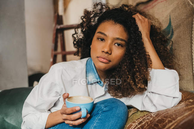 Femme assise sur un canapé avec une tasse de café et regardant la caméra — Photo de stock