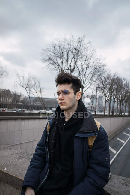 Jeune homme réfléchi dans des lunettes posant sur fond de route asphaltée dans la ville . — Photo de stock