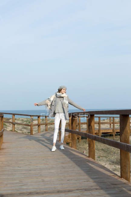Cheerful young stylish woman standing with hands apart on boardwalk at seaside. — Stock Photo