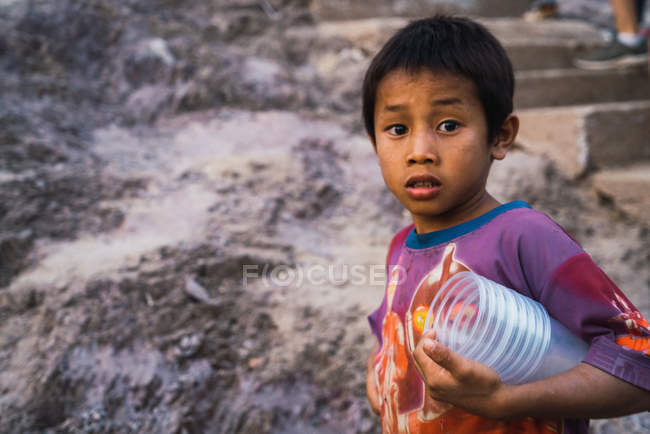 LAOS-FEVEREIRO 18, 2018: Menino com copos de plástico em pé na rocha e olhando para a câmera
. — Fotografia de Stock