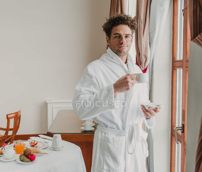 Молодий чоловік в халаті позує з чашкою в готельному номері — стокове фото