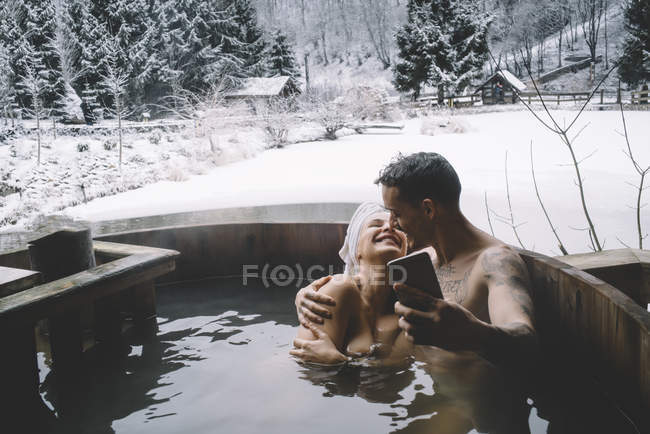 Обнимающая пара, сидящая в ванне на открытом воздухе и делающая селфи — стоковое фото