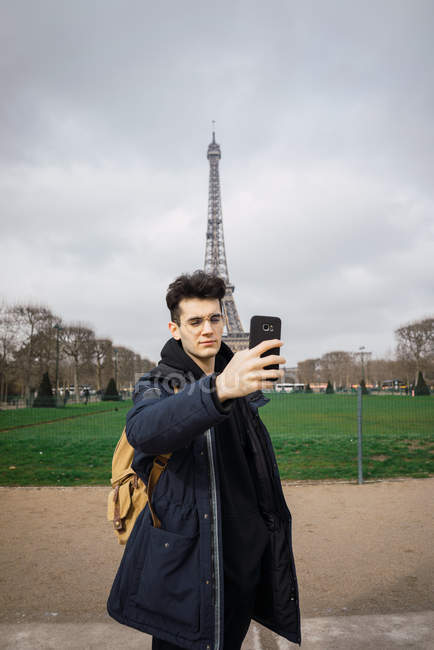 Young man standing with phone and taking selfie on background of Eiffel tower. — Stock Photo