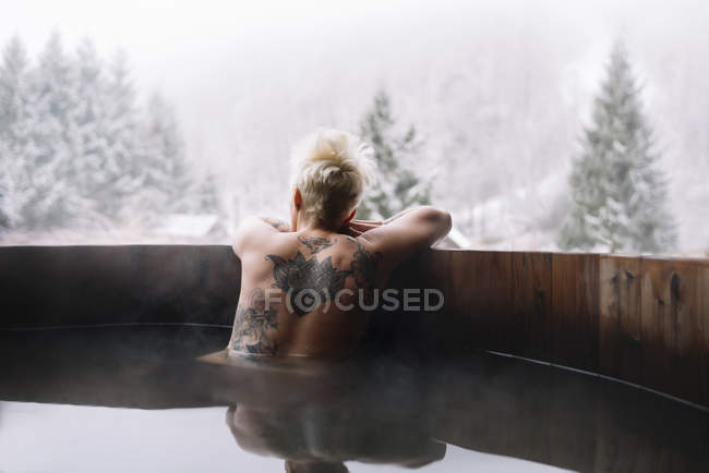 Visão traseira da mulher loira tatuada nadando na banheira de mergulho e admirando a natureza do inverno . — Fotografia de Stock