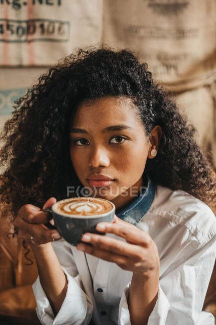 Curly woman drinking cup of coffee and looking at camera — Stock Photo