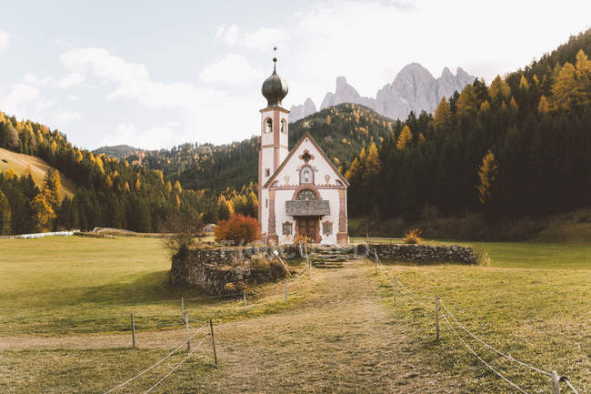 Small white church on green meadow in mountains covered with forest. — Stock Photo