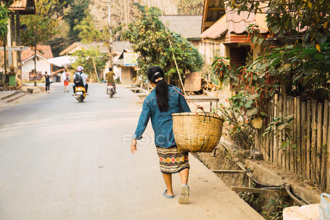 LAOS-FEVEREIRO 18, 2018: Visão traseira da mulher andando na estrada na aldeia e carregando cesta . — Fotografia de Stock