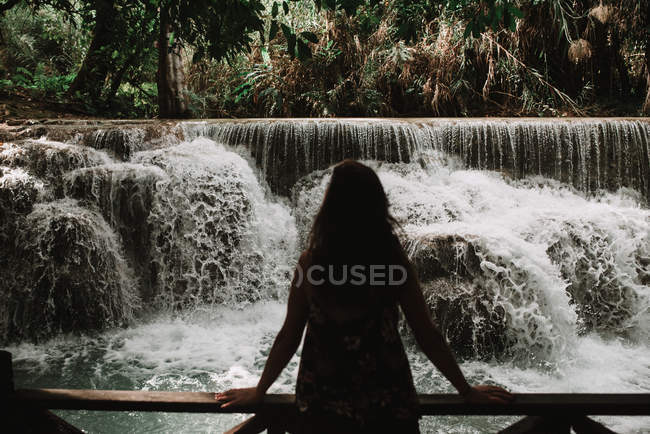 Rückansicht einer Frau, die auf einer Brücke steht und Wasserfall betrachtet. — Stockfoto