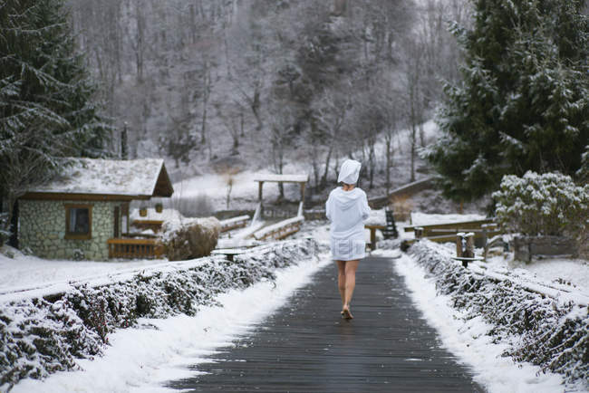 Rear view of woman in bathrobe walking on wooden road to forest in winter. — Stock Photo