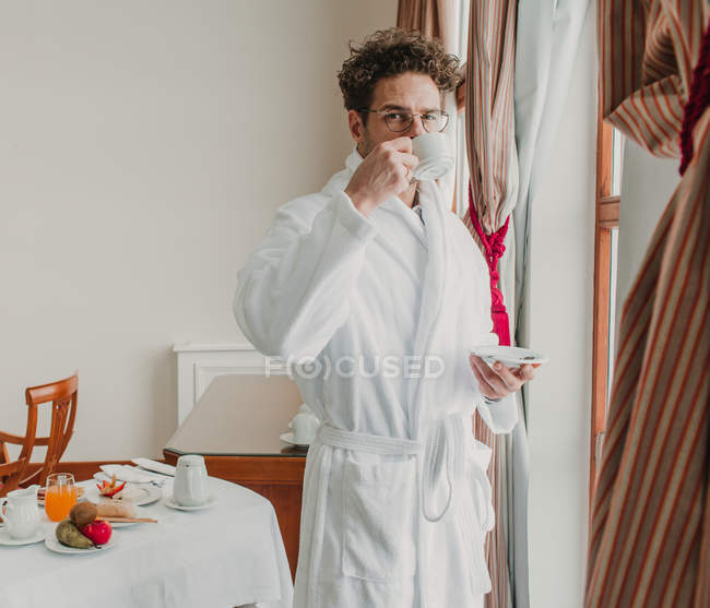 Hombre guapo de pie en el albornoz y beber de la taza de cerámica en la habitación del hotel
. - foto de stock