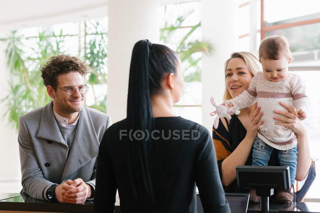 Щаслива сім'я з дитиною розмовляє з жінкою, яка працює на рецепції в готелі — стокове фото