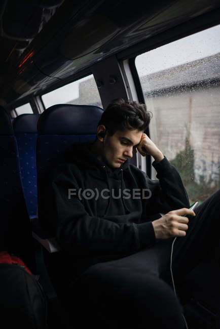 Giovane uomo premuroso seduto e utilizzando smartphone in autobus in giorno di pioggia . — Foto stock