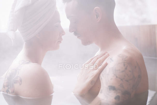 Sensual tattooed couple sitting face to face in plunge tub in winter. — Stock Photo