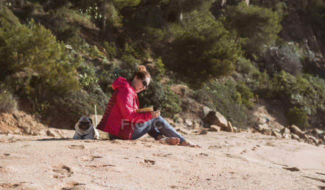 Вид збоку жінки сидить на морському березі і читає книгу і супроводжується собакою — стокове фото