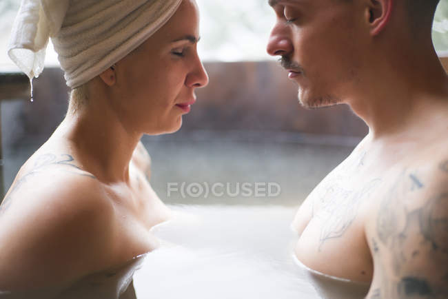 Vista lateral do casal tatuado sensual sentado cara a cara na banheira de mergulho no inverno . — Fotografia de Stock
