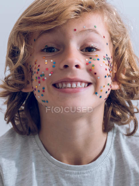 Cheerful young boy with colorful confetti on face looking at came and smiling. — Stock Photo