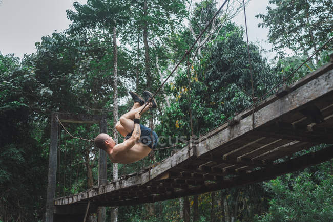 Shirtless acrobatic man hanging on rope of grungy wooden bridge in  forest. — Stock Photo