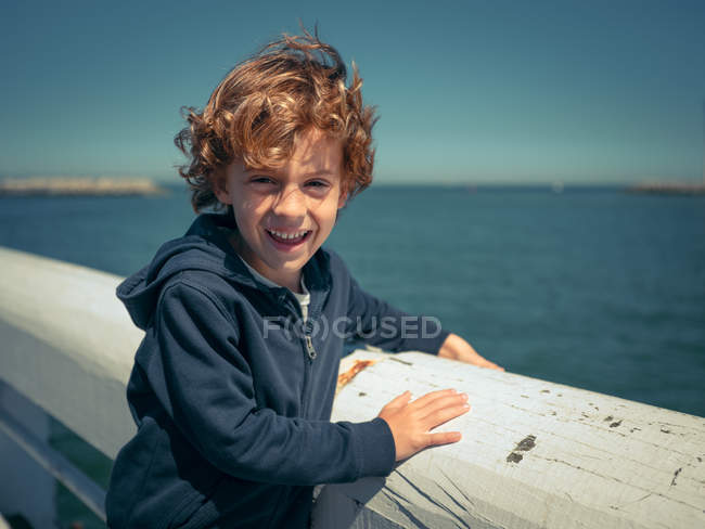 Веселий молодий хлопчик спирається на білий поручень на узбережжі і дивиться на камеру . — стокове фото