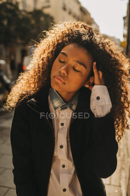 Mulher elegante ajustando o cabelo com os olhos fechados olhos na rua ensolarada — Fotografia de Stock
