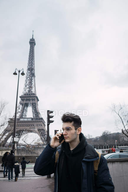 Hombre joven caminando en la calle en el fondo de la torre Eiffel y hablando en el teléfono inteligente - foto de stock