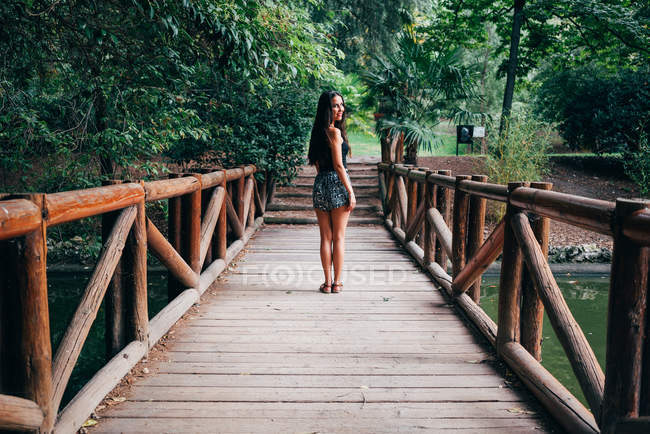 Красивая юная брюнетка стоит на деревянном мосту и смотрит через плечо в камеру
. — стоковое фото