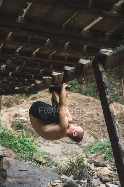Sporty man hanging upside down on wooden bridge in forest. — Stock Photo