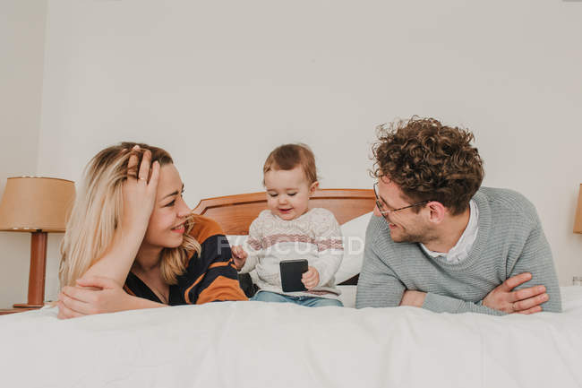 Cheerful couple looking at child with smartphone on bed — Stock Photo