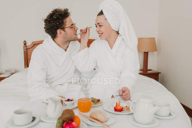 Mujer en albornoz alimentación hombre con desayuno en la cama del hotel - foto de stock
