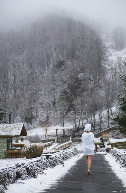 Visão traseira da mulher em roupão de banho andando na estrada de madeira no resort de inverno — Fotografia de Stock