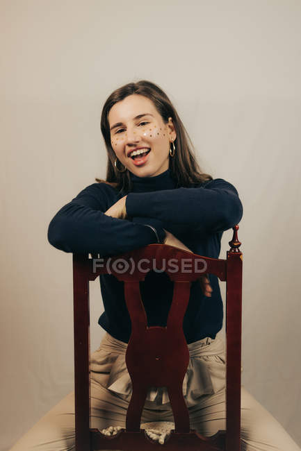 Smiling woman with glitters on face sitting on wooden chair and looking at camera — Stock Photo