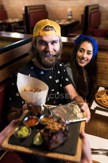 Sonriente hombre barbudo sentado con su novia en la cafetería y tomando comida de camarero
. - foto de stock