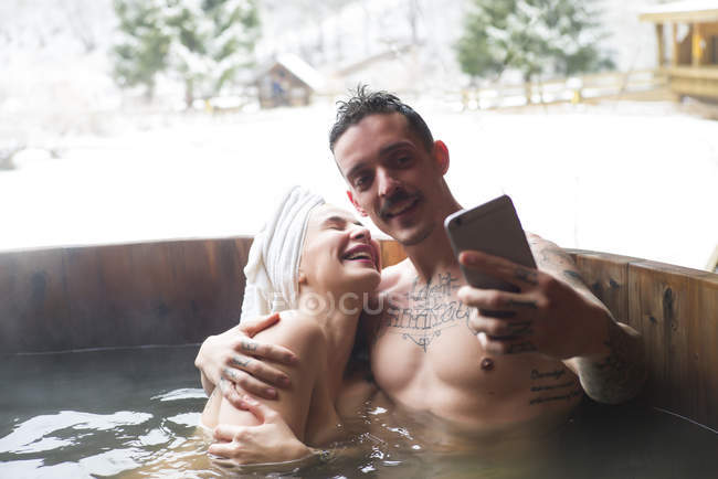 Sensual pareja tatuada sentada en una bañera profunda y tomando selfie - foto de stock