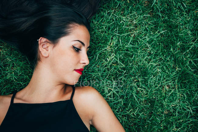 Брюнетка с красными губами лежит в траве и смотрит в сторону — стоковое фото