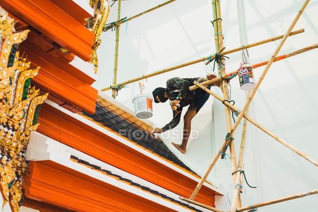CHIANG RAI, THAILAND- FEBRUARY 12, 2018: Decorator man hanging on construction site and painting roof of Asian building. — Stock Photo