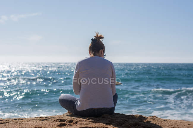 Задний вид женщины, сидящей на берегу моря весной и читающей книгу — стоковое фото