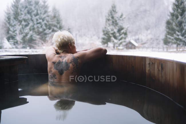 Visão traseira da mulher loira tatuada relaxando na banheira de mergulho e admirando a natureza do inverno . — Fotografia de Stock