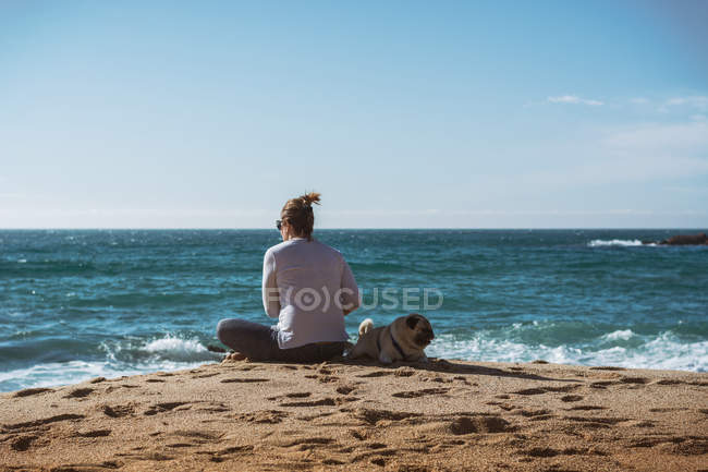 Femme mûre assise sur le bord de la mer avec chiot de carlin de côté — Photo de stock