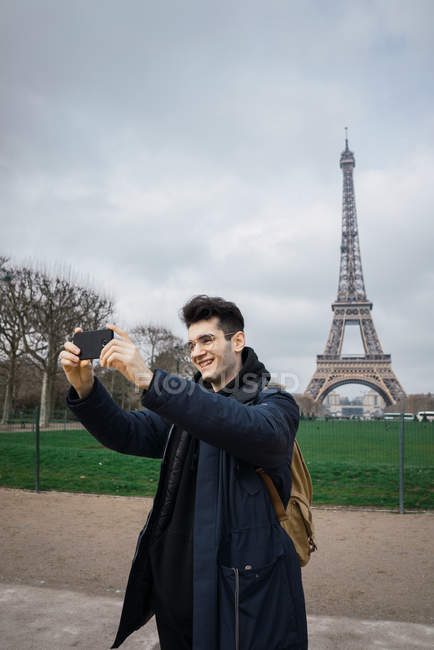 Joven parado con el teléfono y tomando selfie en el fondo de la torre Eiffel . - foto de stock
