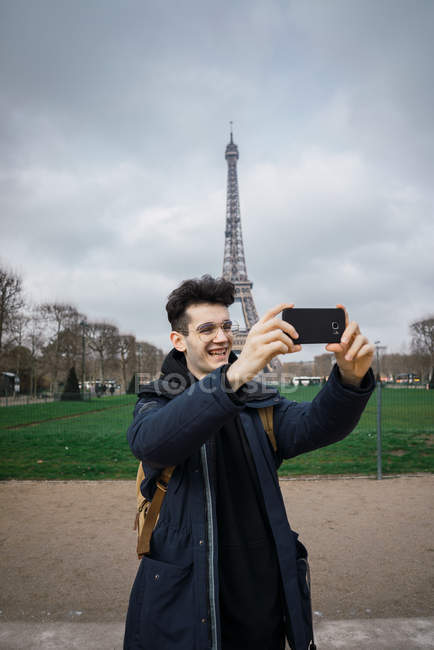Молодой человек в очках стоит с телефоном и делает селфи на фоне Эйфелевой башни . — стоковое фото