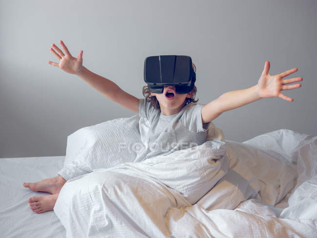 Adorable little boy with hands apart sitting on bed and playing with VR headset. — Stock Photo