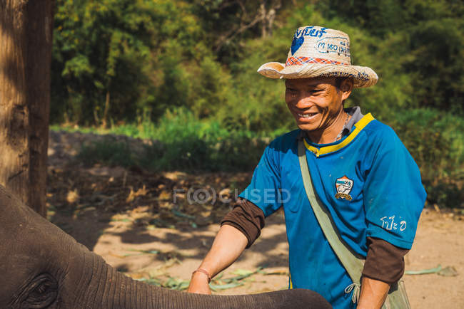 CHIANG RAI, THAILAND- FEBRUARY 12, 2018: Cheerful man standing in nature and stroking baby elephant. — Stock Photo