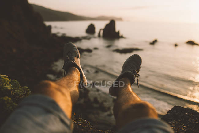 Gambe dell'uomo seduto sul bordo della scogliera al mare . — Foto stock