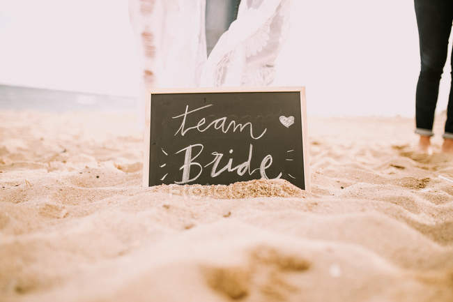 Tableau noir avec lettrage de mariée d'équipe dans le sable et les jambes de culture des personnes . — Photo de stock