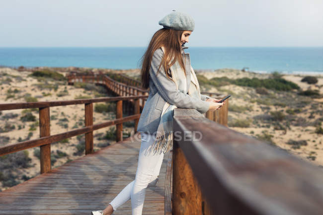 Cheerful woman browsing smartphone on boardwalk at sunny seaside — Stock Photo